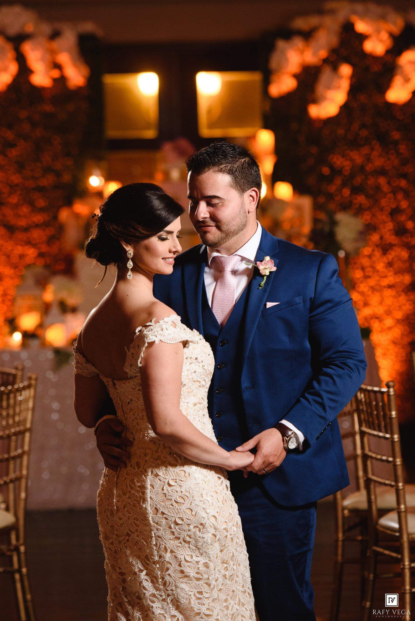 Bride in lace gown and groom in navy suit share romantic moment at elegantly lit wedding reception venue.