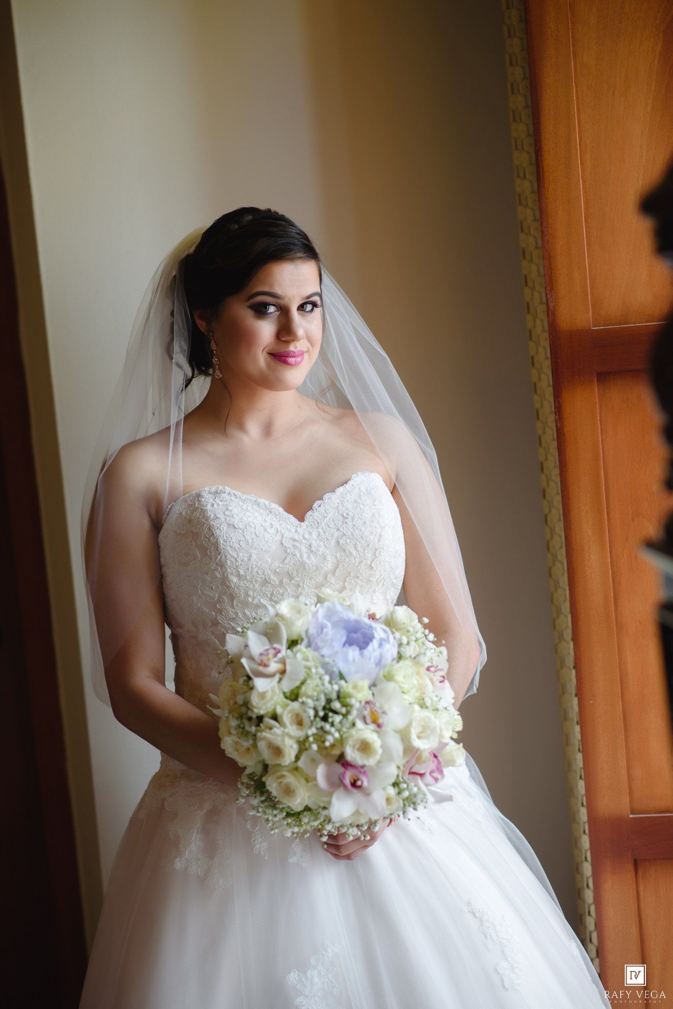 Bride in strapless lace ballgown holding white and lavender bouquet with veil, posing indoors near window.