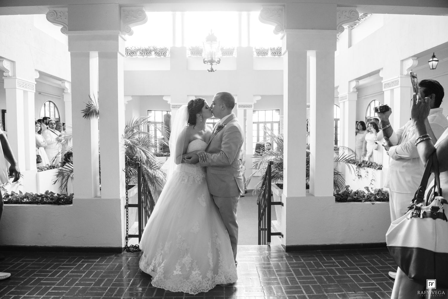 Bride and groom share romantic first kiss in elegant venue aisle surrounded by guests in black and white photo.