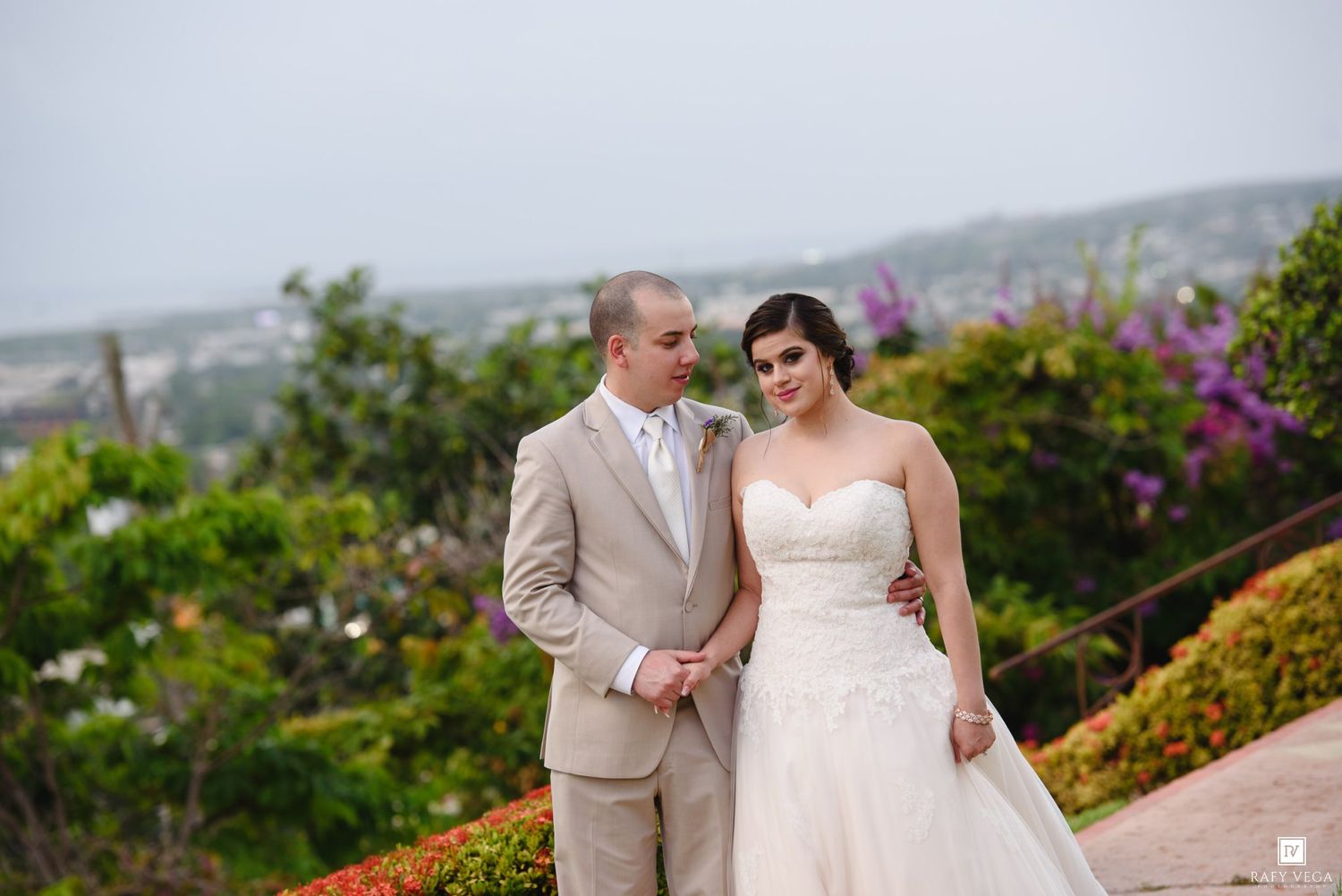 Bride in white strapless gown and groom in tan suit pose together outdoors with lush greenery and cityscape backdrop.