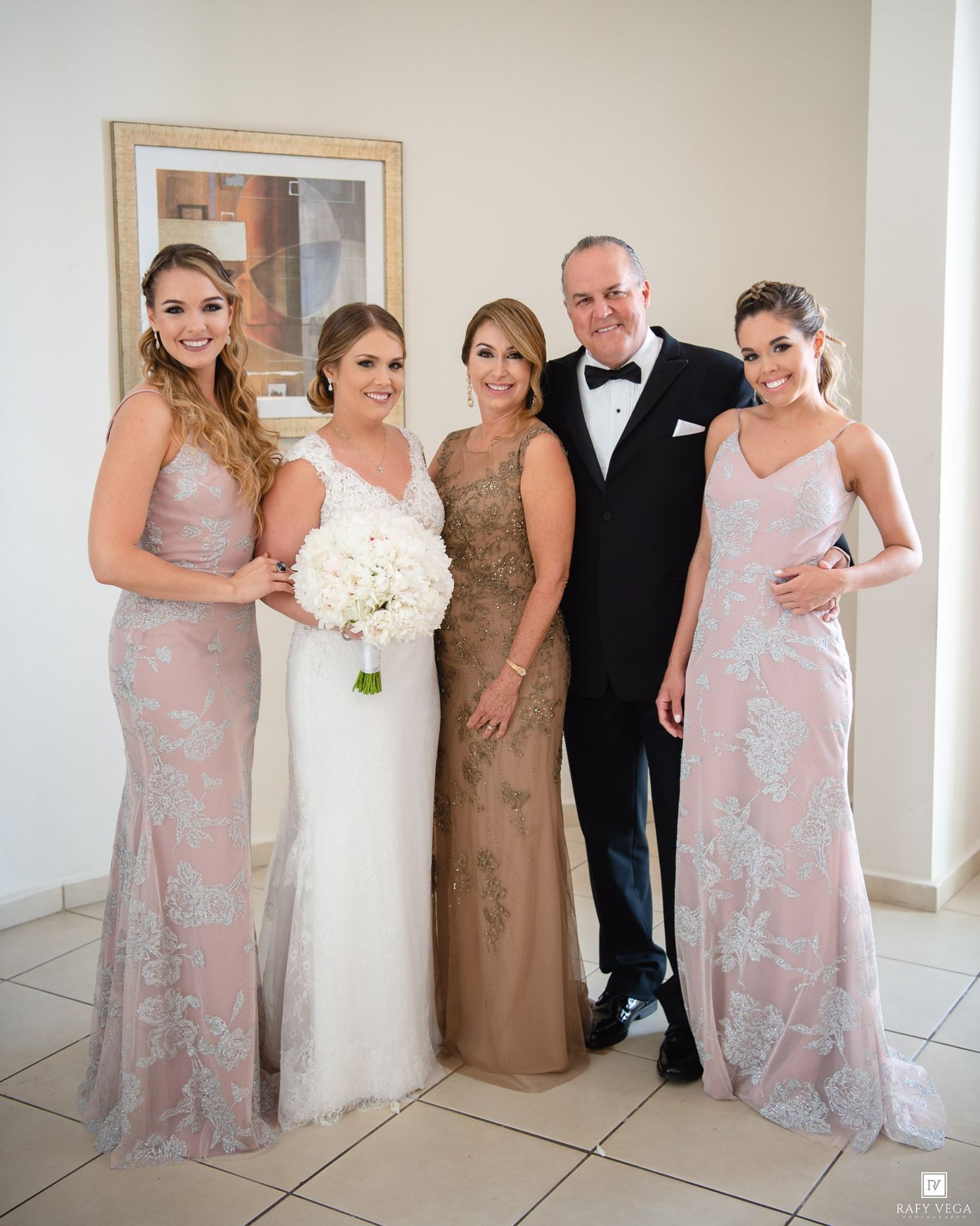 Wedding party poses together, bride in white gown holding bouquet, flanked by bridesmaids in pink lace dresses and parents in formal attire.
