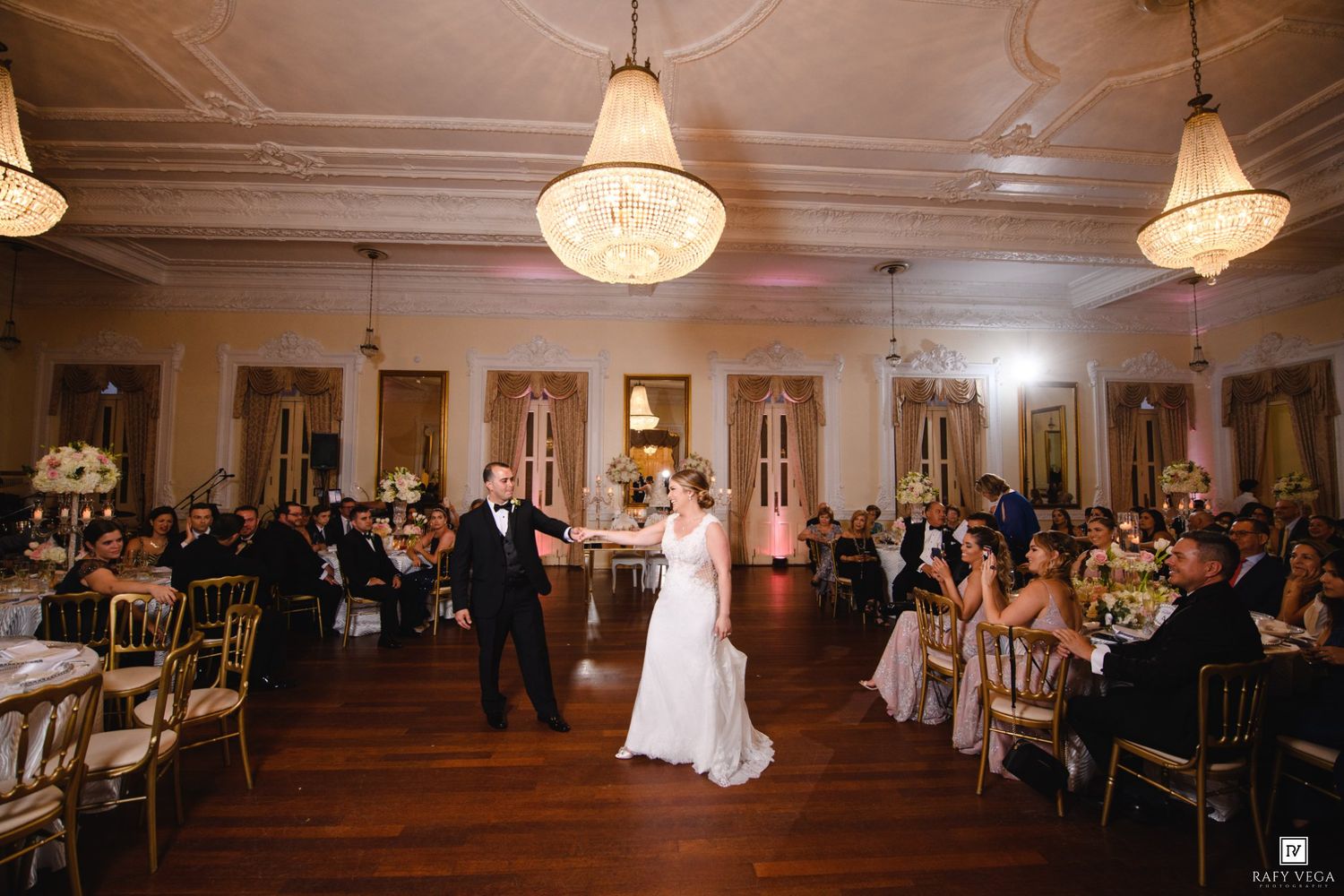Bride and groom share first dance in elegant ballroom with crystal chandeliers and gold chairs at wedding reception.