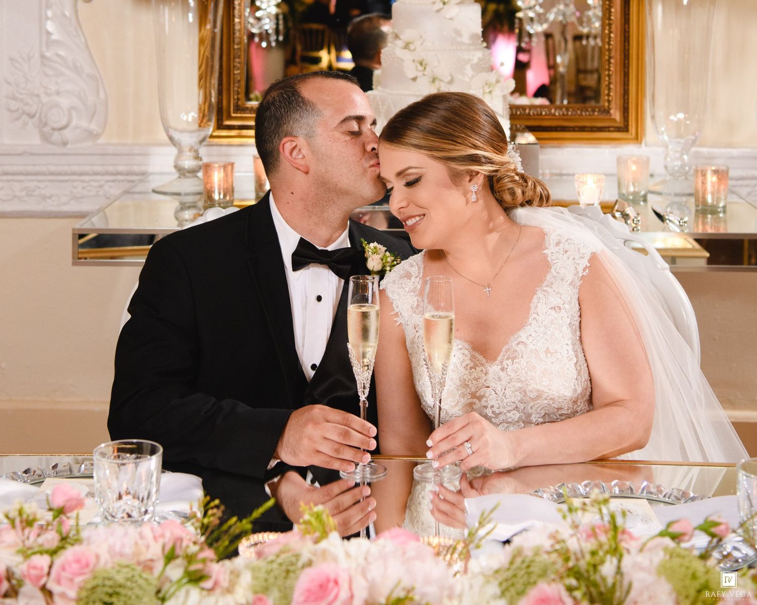 Bride and groom share a kiss while holding champagne glasses at elegant wedding reception with floral centerpieces.