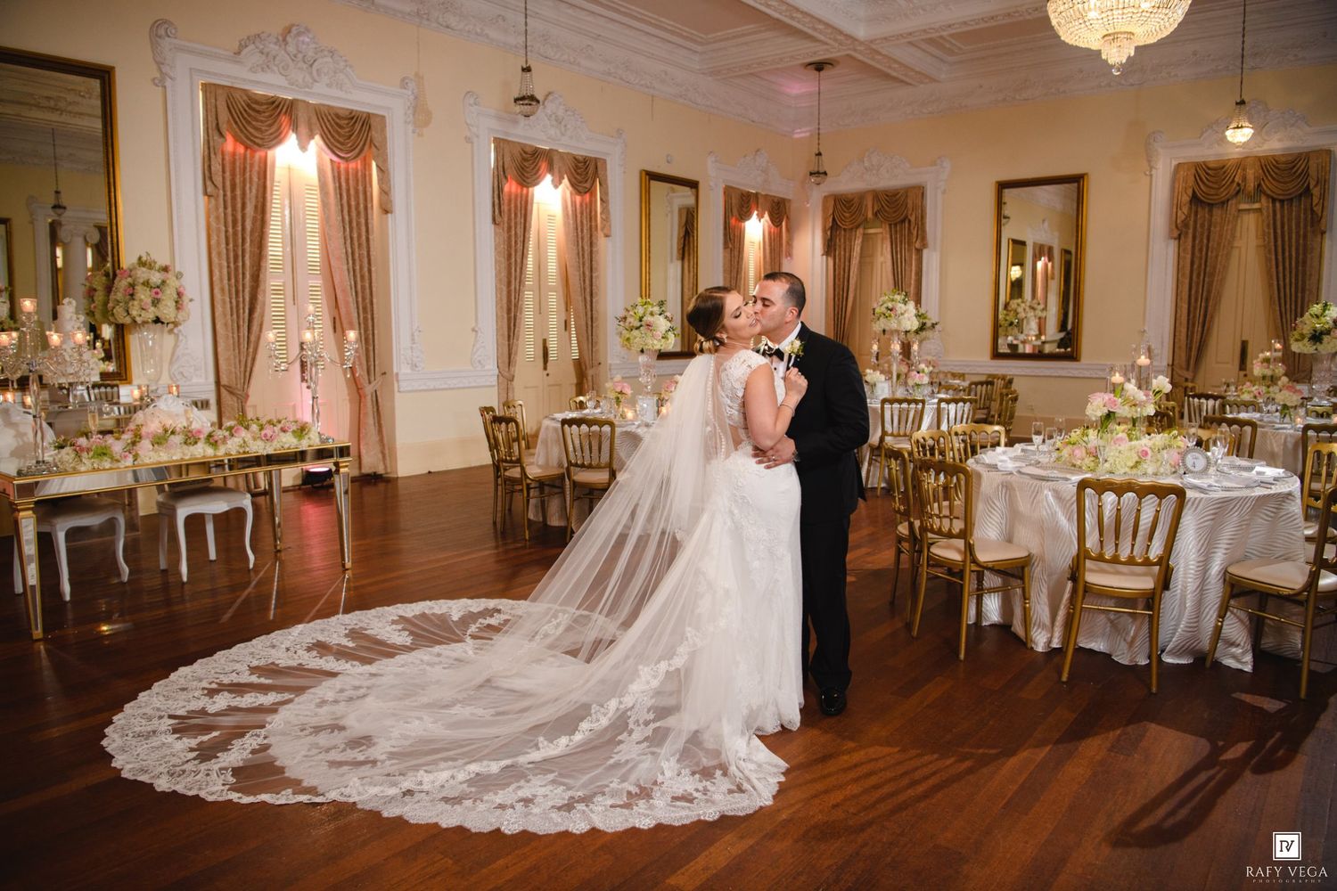 Bride and groom sharing first kiss in ornate gold and white wedding reception ballroom with elegant decor.
