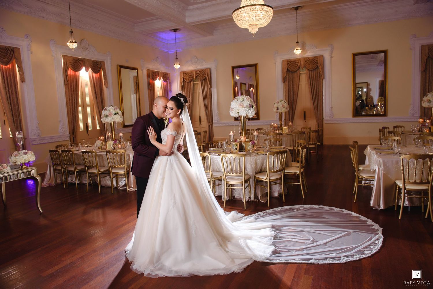 Bride and groom share romantic embrace on dance floor of elegantly decorated wedding reception venue with gold chairs.