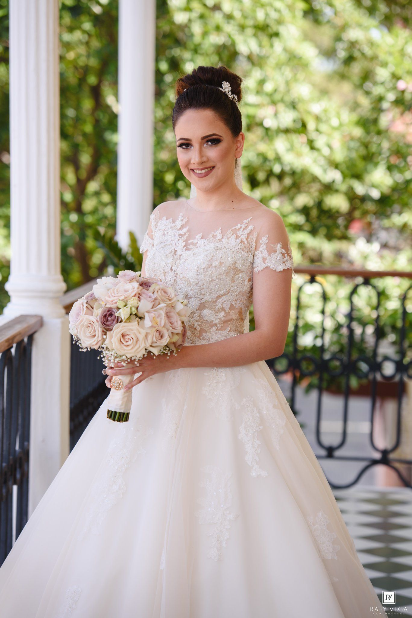 Smiling bride in lace ball gown wedding dress holding pink rose bouquet on elegant venue balcony.