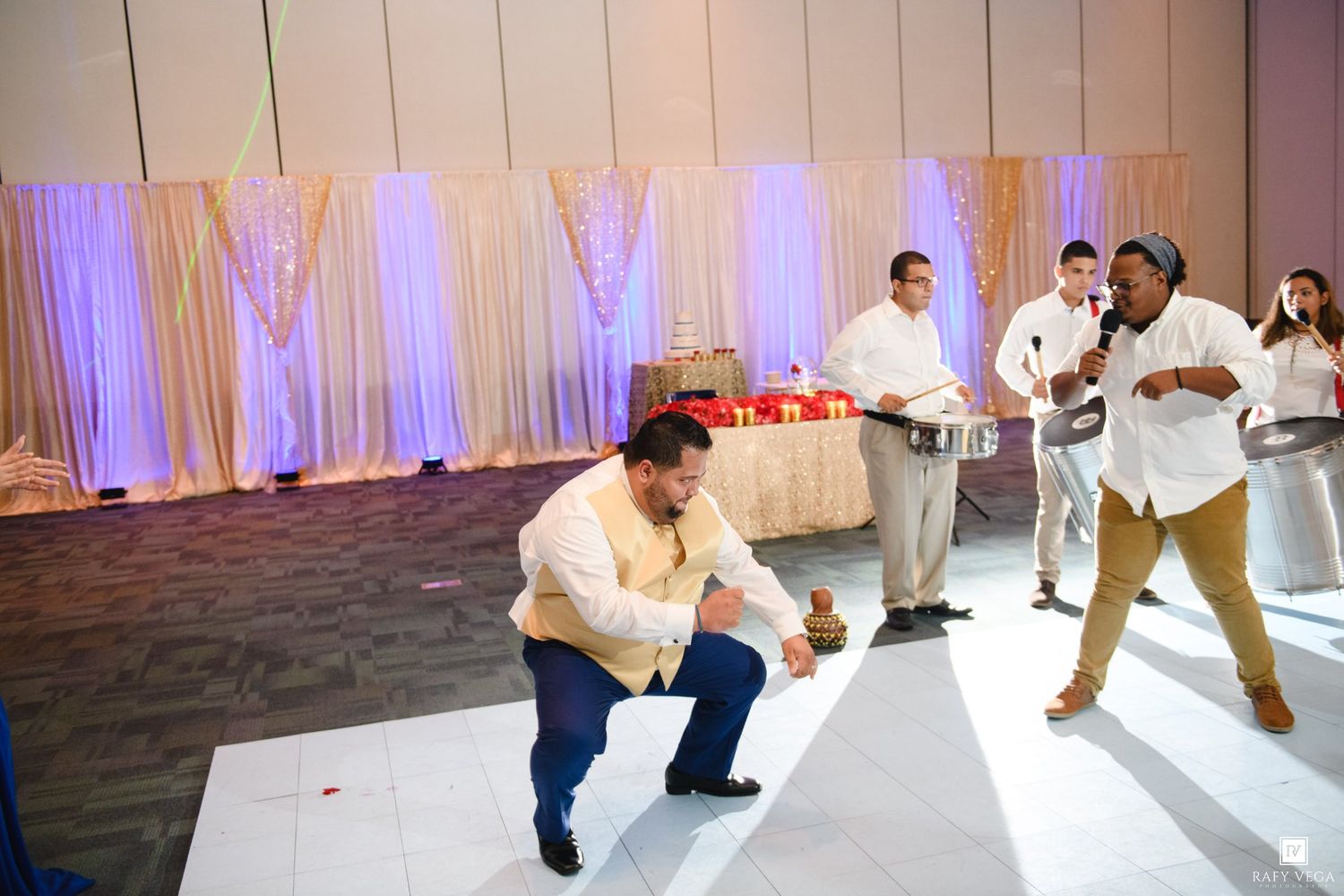Man crouching on dance floor at elegant wedding reception with decorative backdrop and guests nearby.