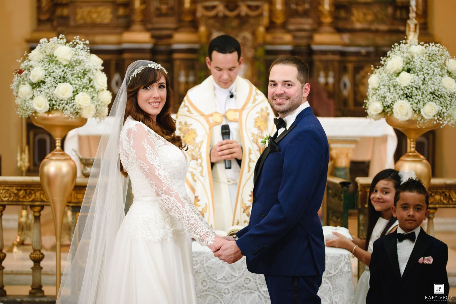 Bride and groom holding hands at church altar during wedding ceremony with priest and children in background.
