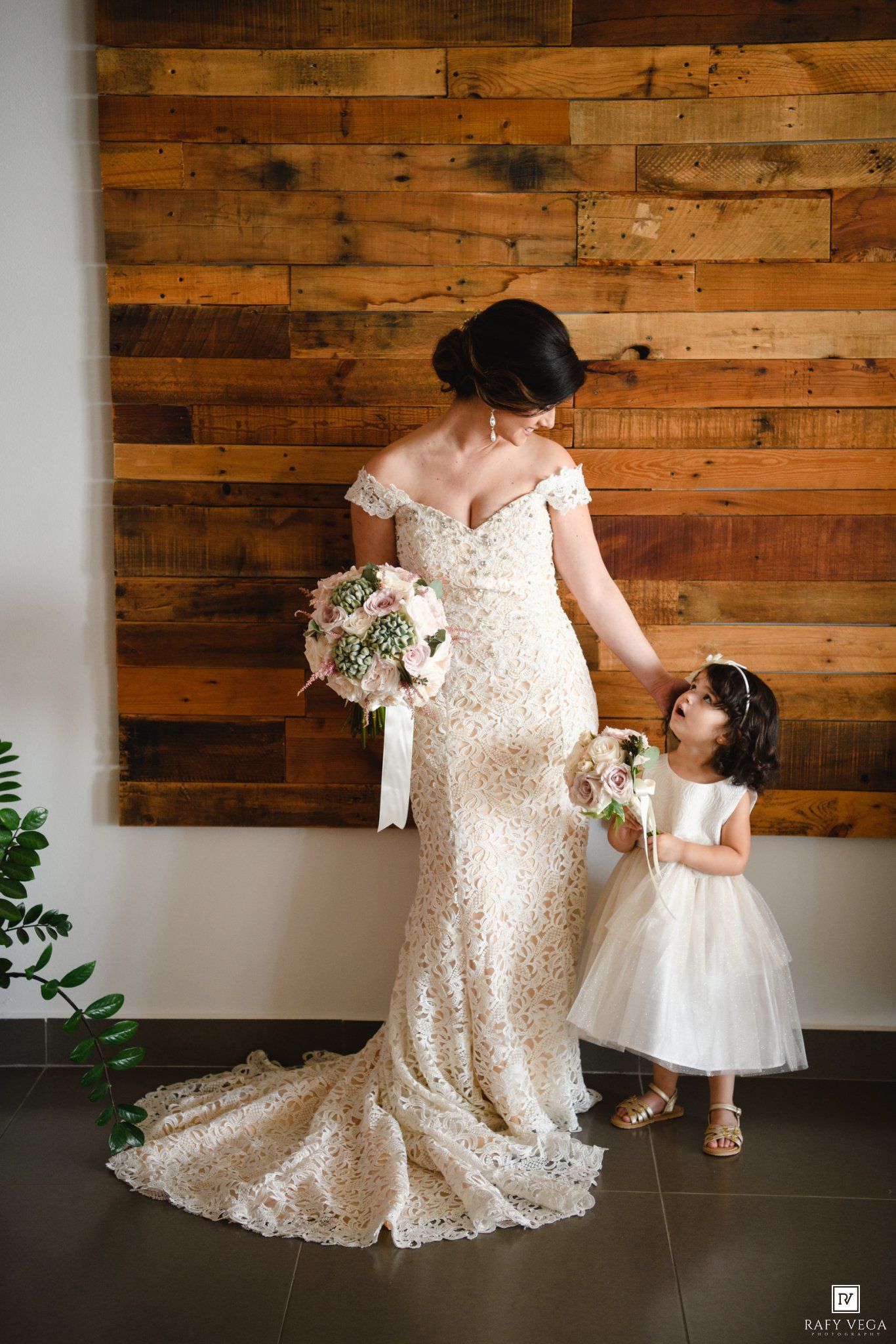 Bride in lace gown holds bouquet beside flower girl in white dress against rustic wood plank wall.