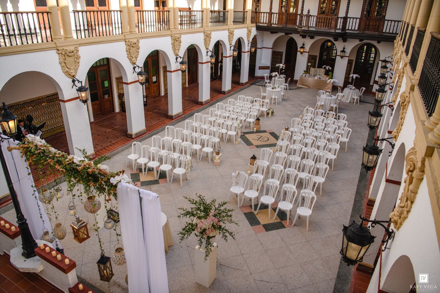 Elegant outdoor courtyard wedding setup with white chairs, floral arrangements, and arched colonial architecture.