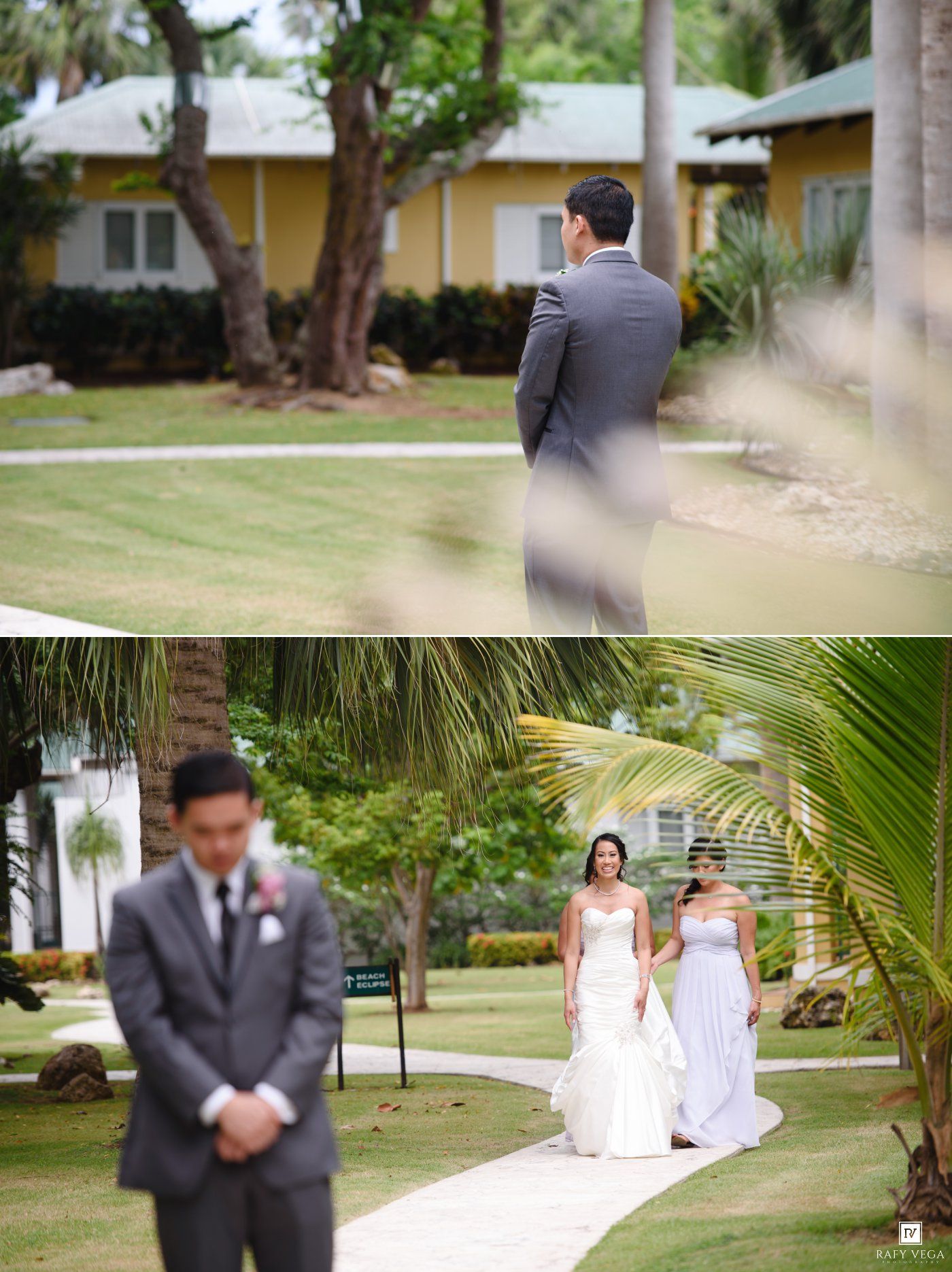 Groom in gray suit awaits bride walking down outdoor path with bridesmaid at tropical wedding venue.