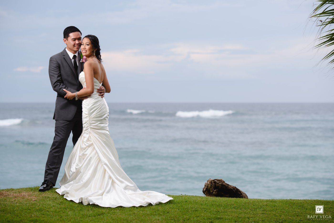 Bride and groom embrace in wedding attire on a lush green lawn with ocean waves in the background.