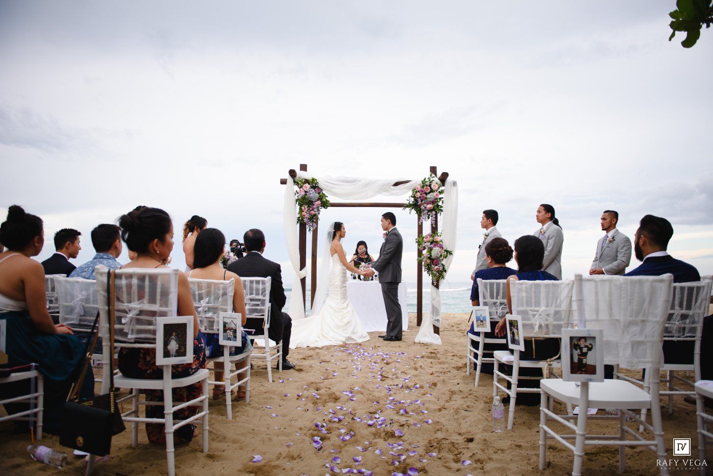 Elegant beach wedding ceremony with white chairs, floral arch, and guests seated by the ocean.