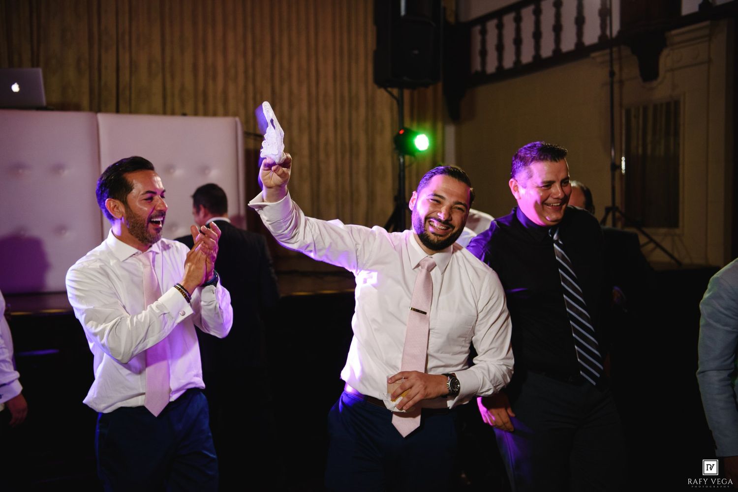Men in dress shirts celebrate joyfully at a wedding reception, one raising his hand triumphantly on the dance floor.