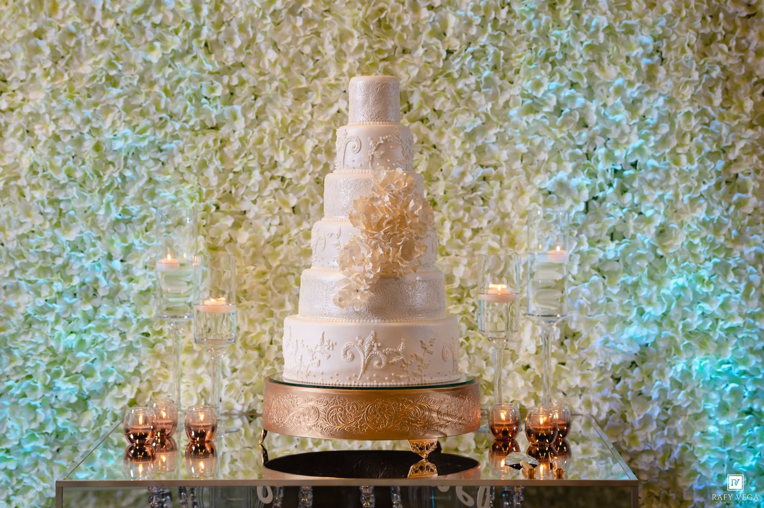 Elegant multi-tier white and gold wedding cake displayed against a lush floral wall with candles.