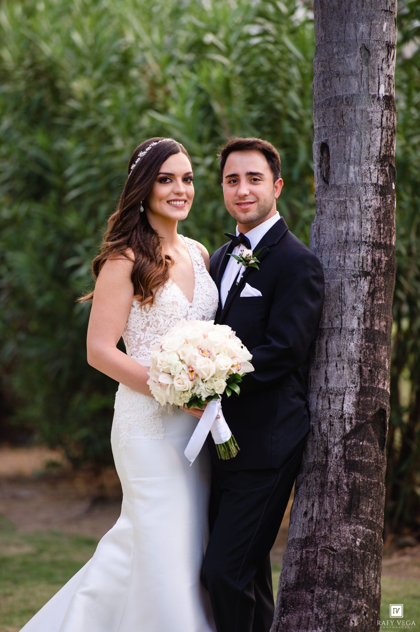 Bride in fitted lace wedding gown holding white bouquet poses with groom in black tuxedo beside a palm tree.