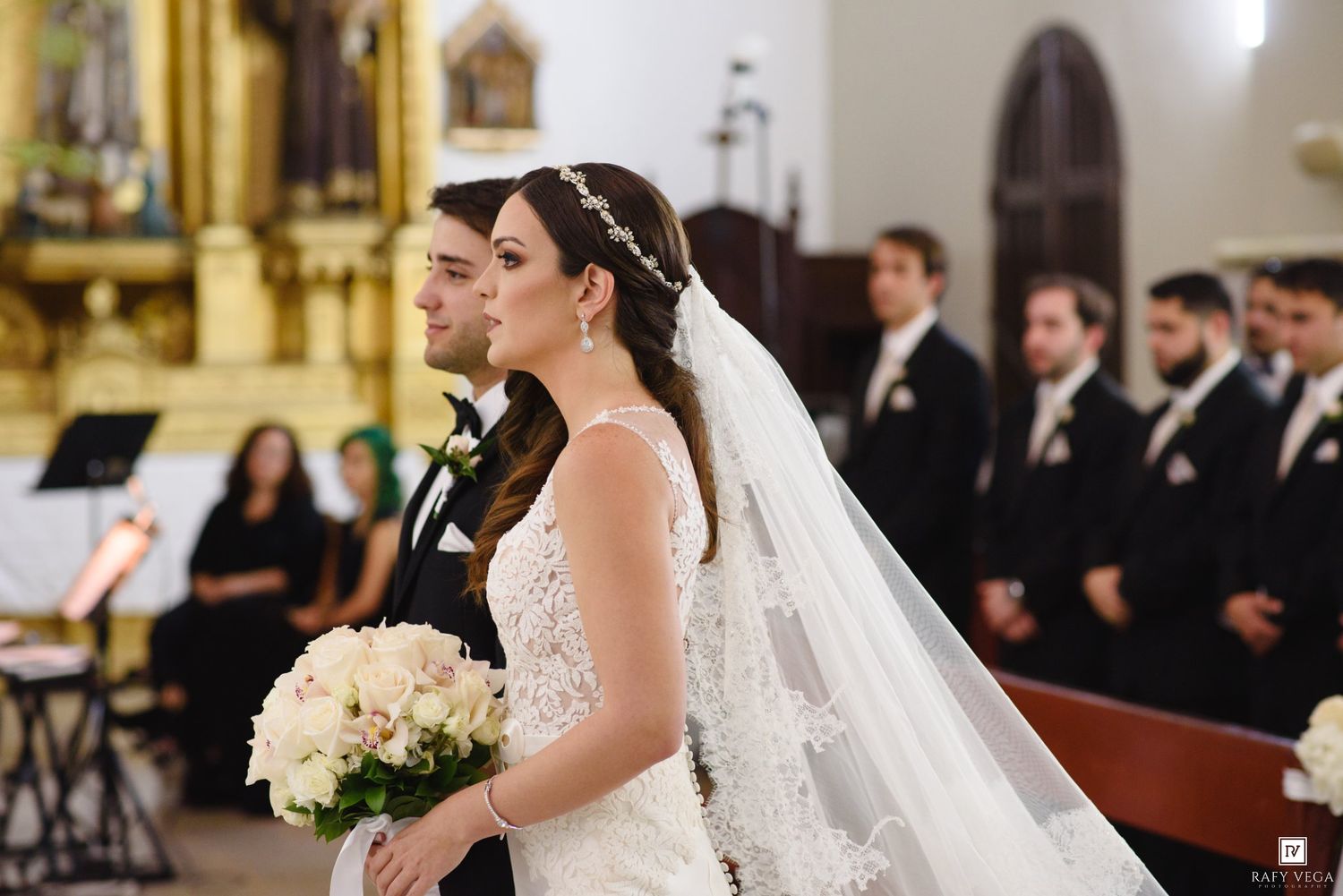 Bride in lace gown and veil holds white bouquet beside groom in church ceremony aisle with wedding party.