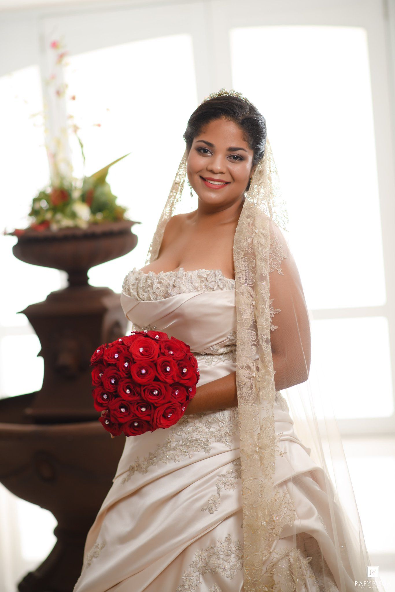 Smiling bride in strapless white gown with lace veil holding red rose bouquet near decorative fountain.