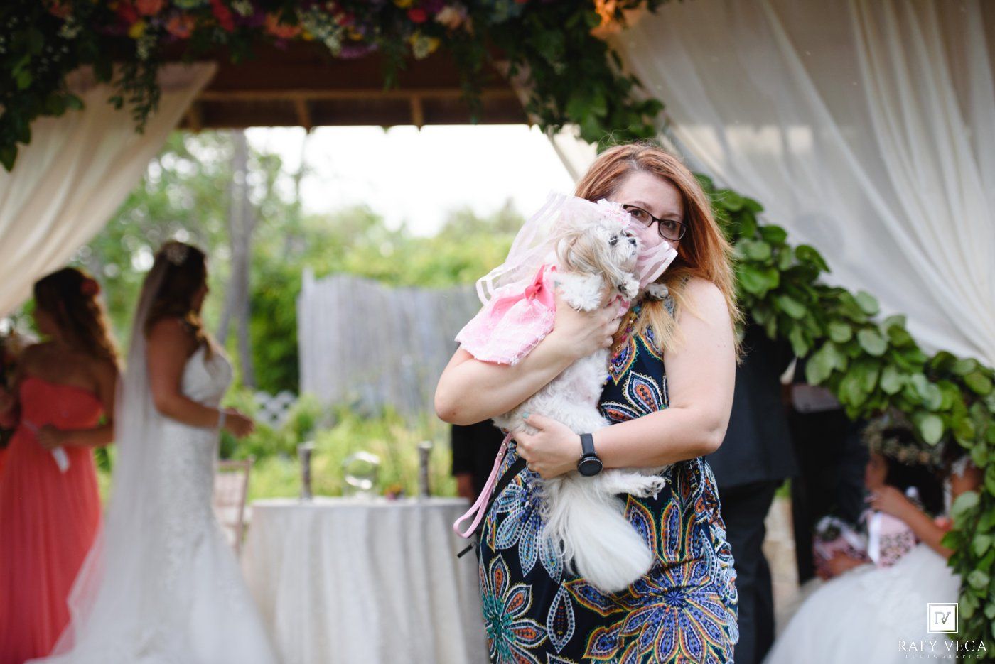 Woman in blue dress holds small dog dressed in pink at an outdoor wedding ceremony.