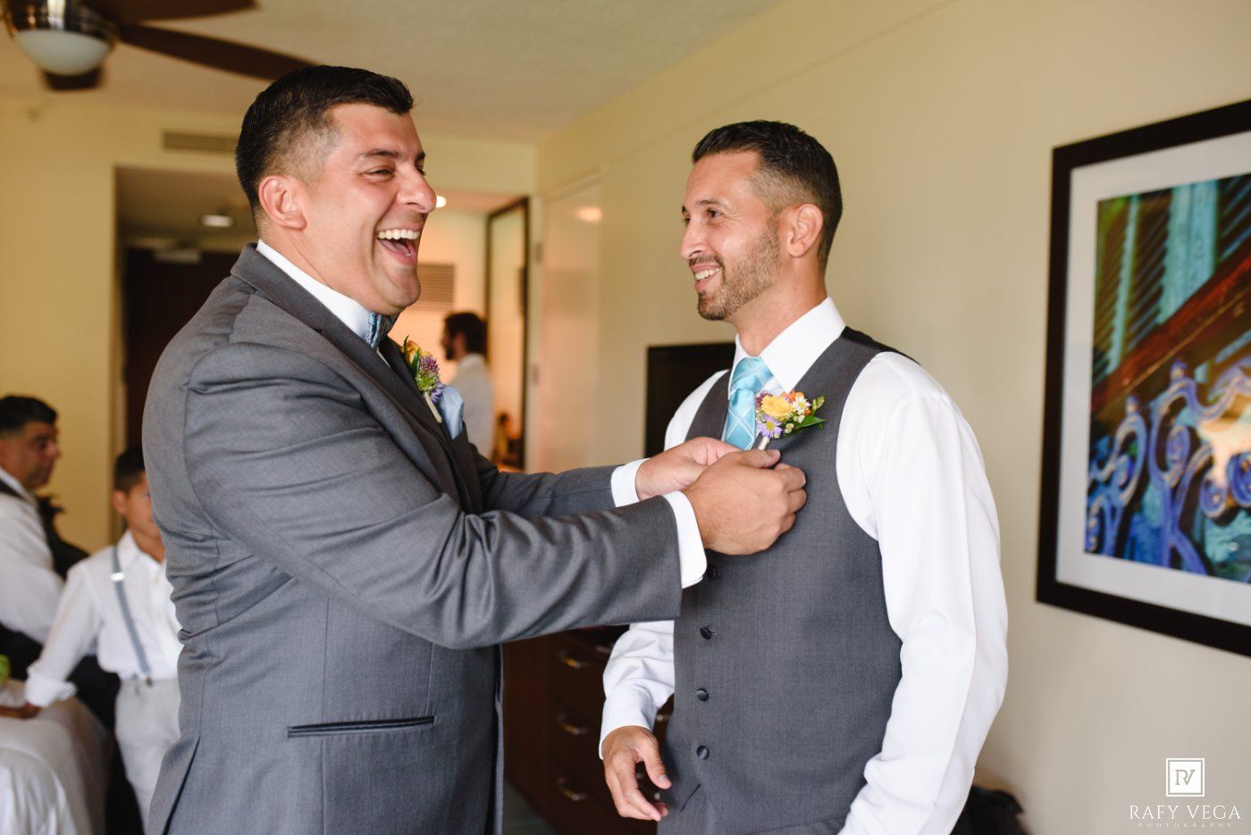 Groom laughing with groomsman while getting boutonniere pinned on vest during wedding preparations indoors.