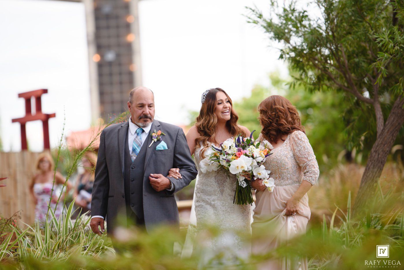 Bride walks down outdoor aisle with parents, holding floral bouquet, surrounded by lush greenery at wedding ceremony.