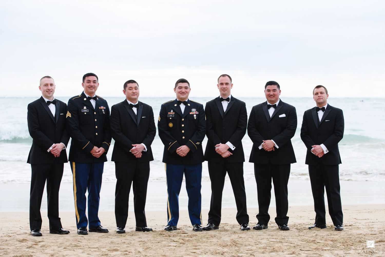 Military groomsmen in dress uniforms pose with groom on beach for wedding photo.