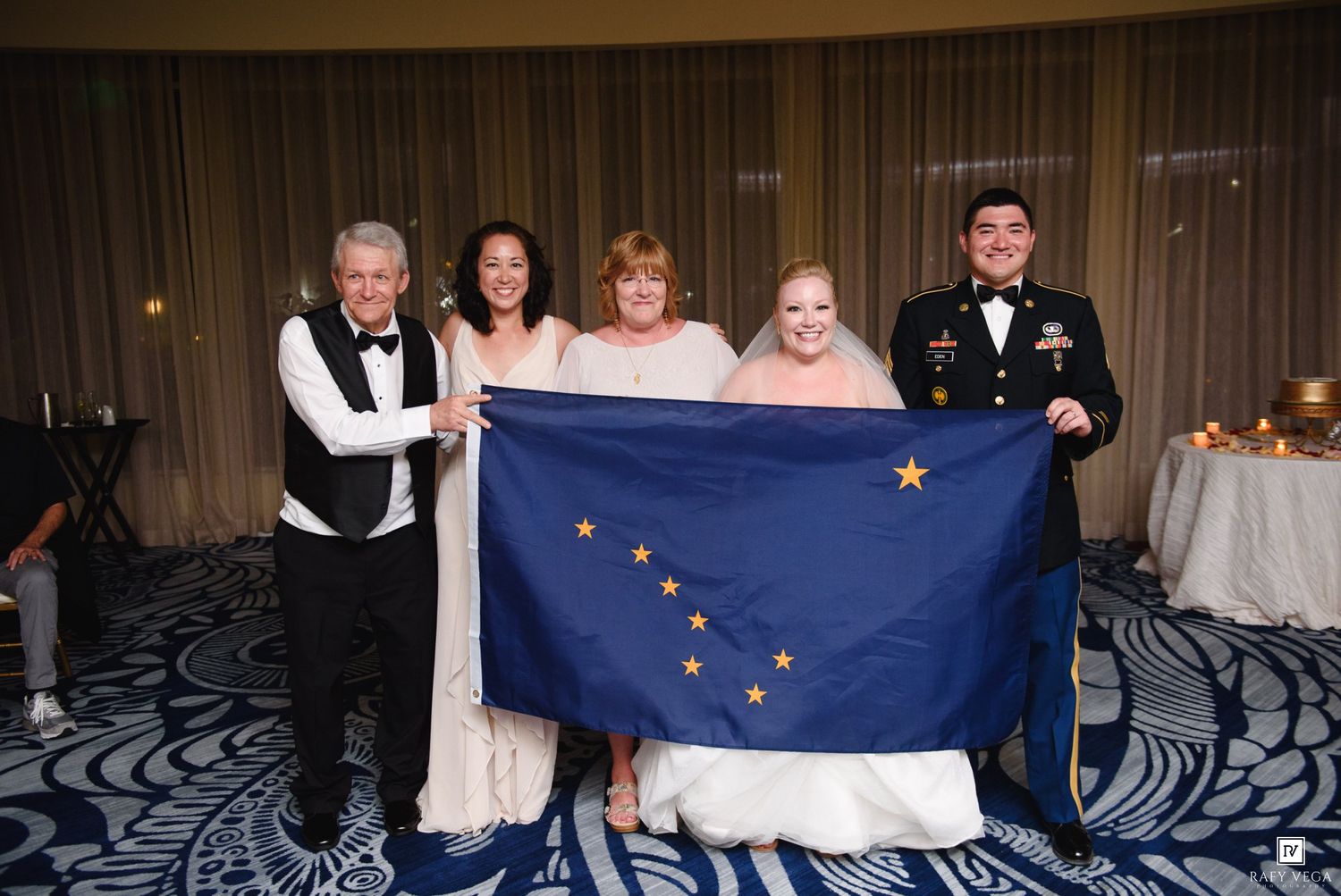 Group of five people smiling and holding an Alaska state flag at a formal indoor event.