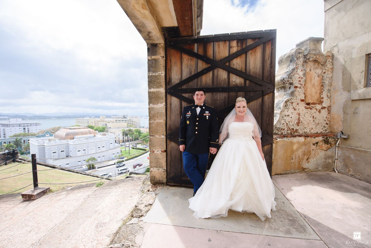Military groom and bride in white gown exit historic stone doorway with scenic city view backdrop.