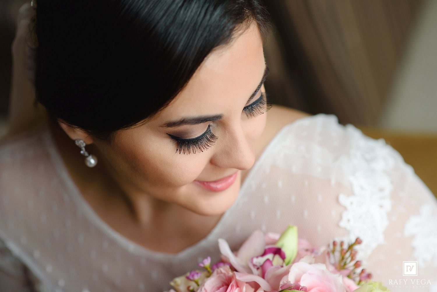 Elegant bride with dark hair, pearl earrings, and pink floral bouquet wearing a lace wedding dress on her wedding day.