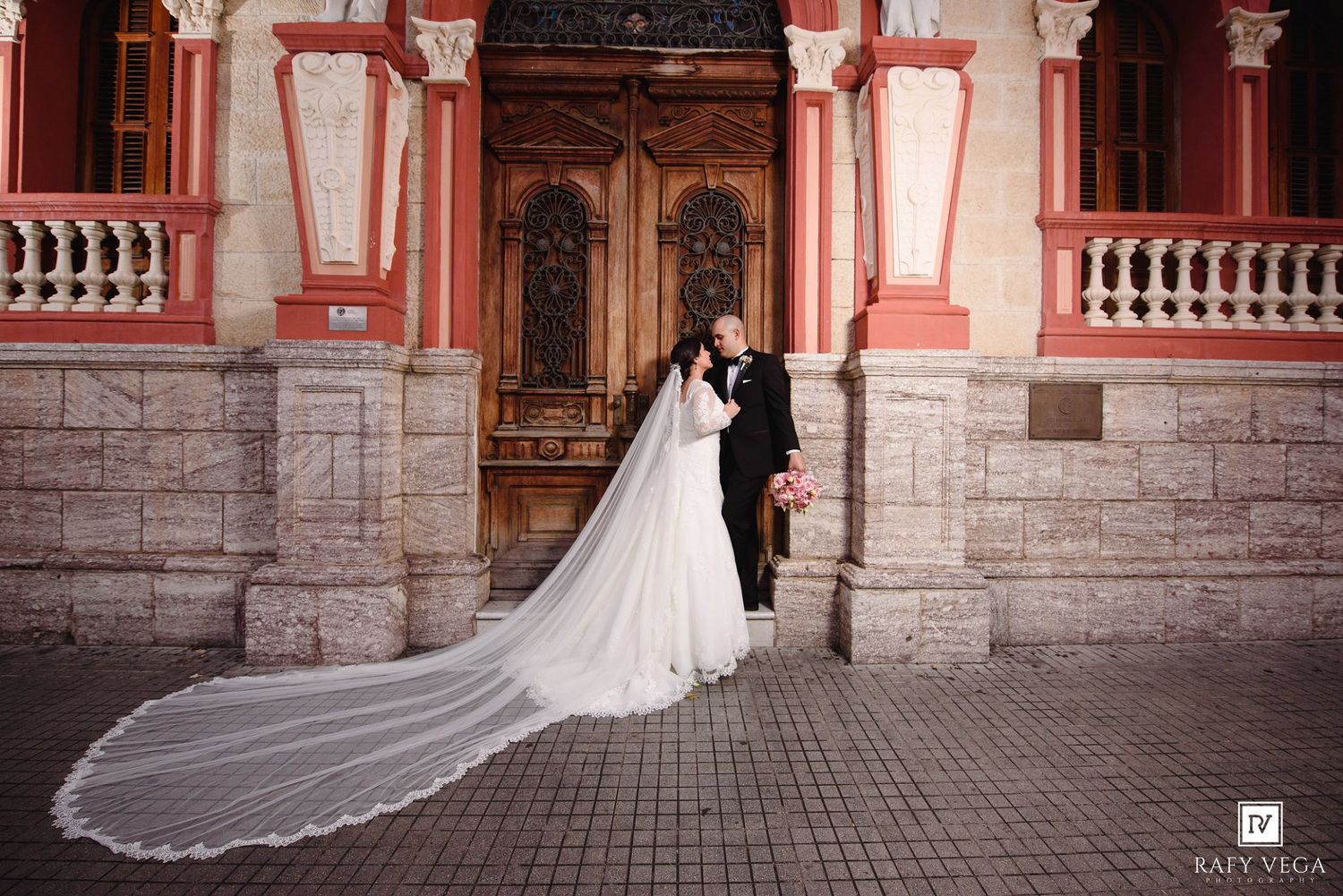 Bride and groom share a romantic kiss outside a historic red building, her long cathedral veil trailing elegantly on the ground.