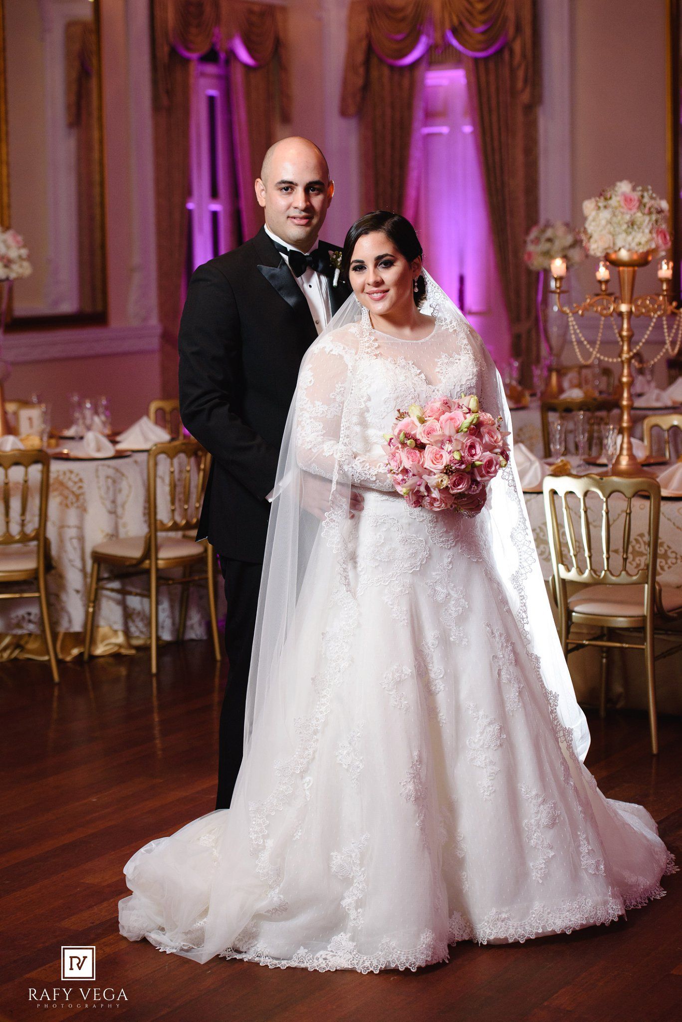Bride and groom posing in elegant ballroom, bride in lace gown with veil holding pink bouquet.