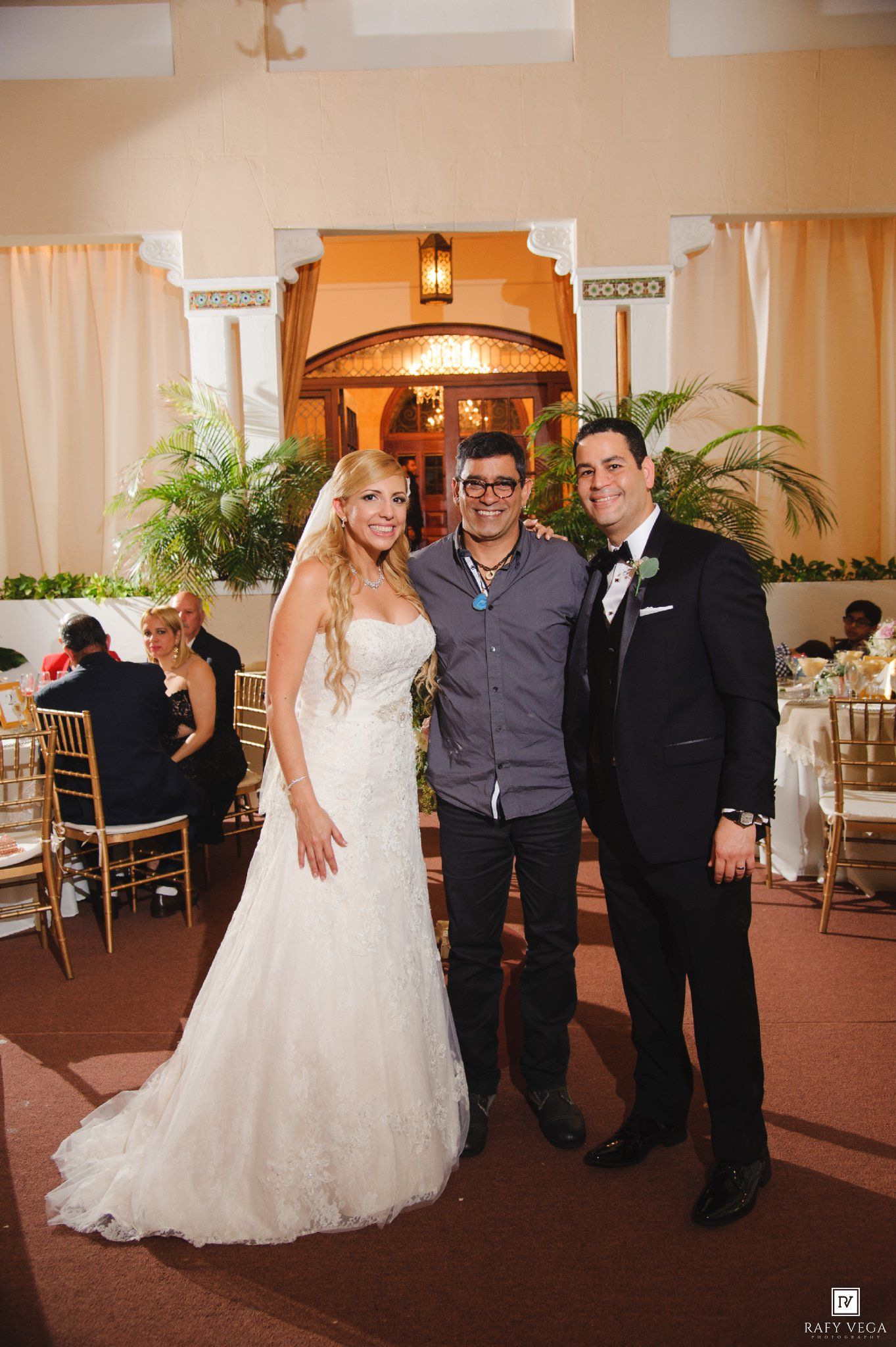 Bride in white gown and groom in tuxedo pose with guest at elegant wedding reception venue.