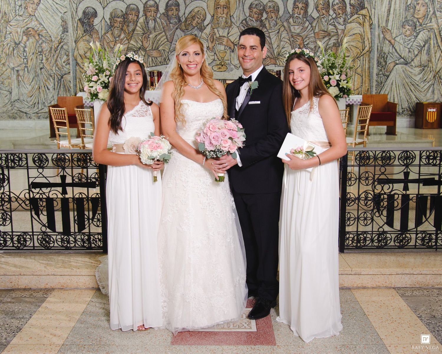 Bride and groom pose with two bridesmaids in white dresses holding pink bouquets inside an elegant church.