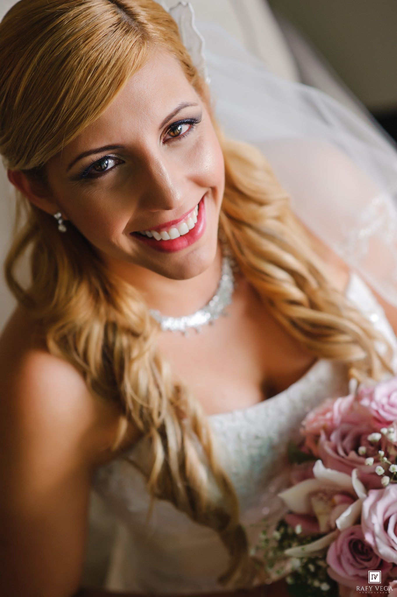 Smiling bride with blonde hair, veil, pearl necklace, and pink rose bouquet in a white strapless wedding gown.