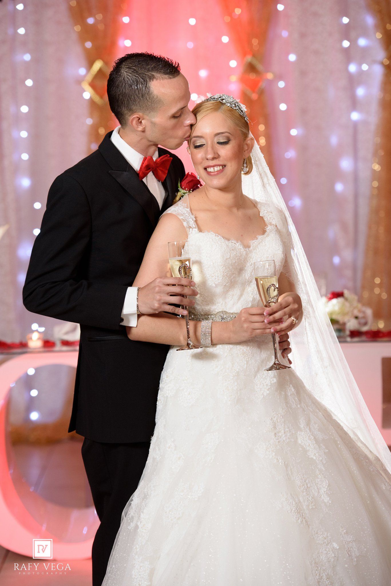 Bride and groom share a champagne toast, groom kissing bride on cheek, wearing wedding attire at elegant reception.