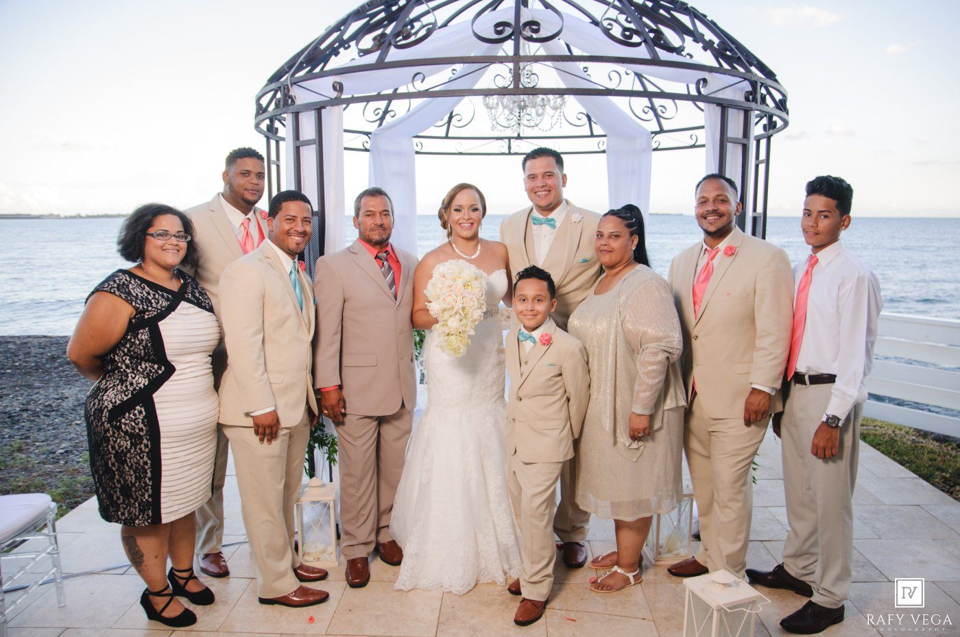 Wedding party poses near oceanside gazebo, bride in white gown with bouquet, groomsmen in tan suits with coral ties.