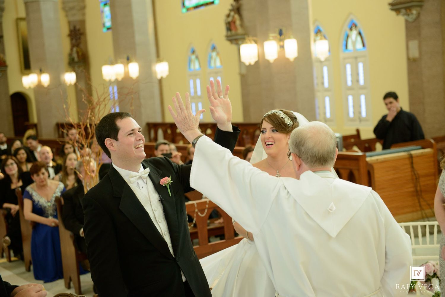 Bride and groom celebrate at church altar with officiant raising hands during wedding ceremony.