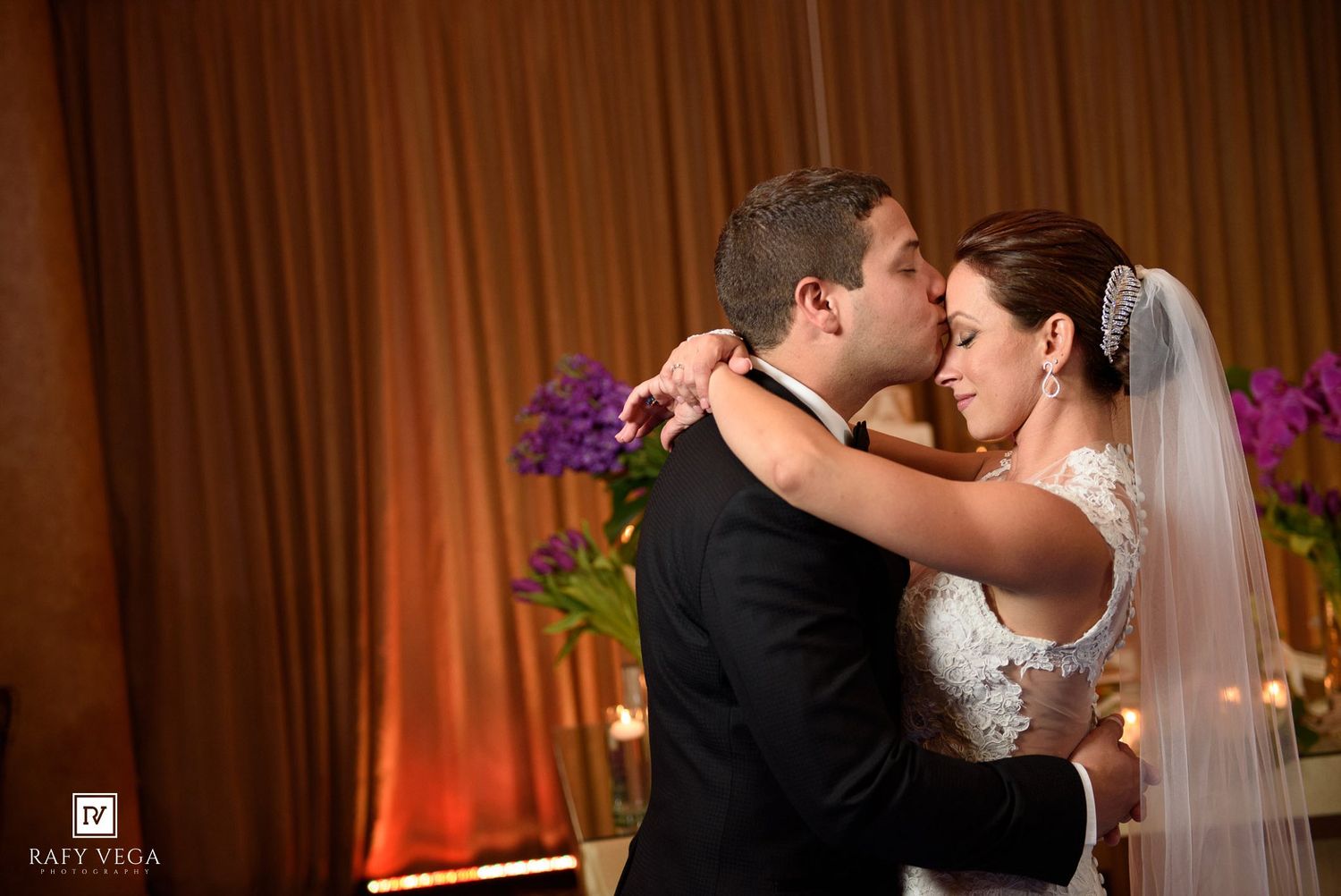 Bride and groom share an intimate first dance embrace, with soft candlelight and purple floral arrangements in the background.