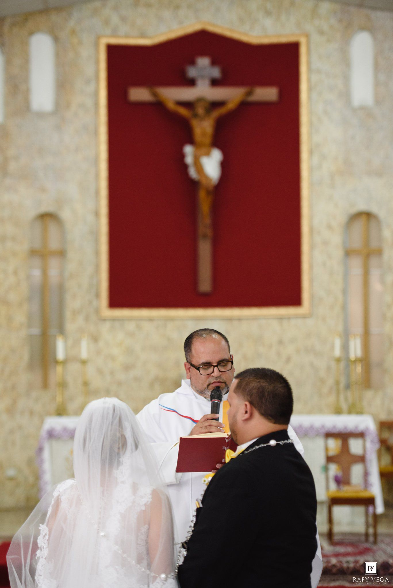 A priest officiates a Catholic wedding ceremony at the altar with a crucifix, as the bride and groom stand before him.