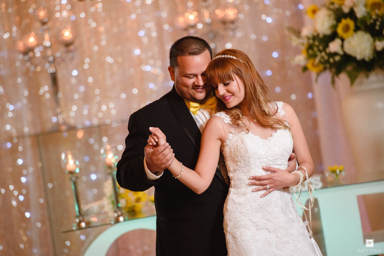 A bride and groom share their first dance, smiling tenderly, with sparkling lights and floral decorations in the background.