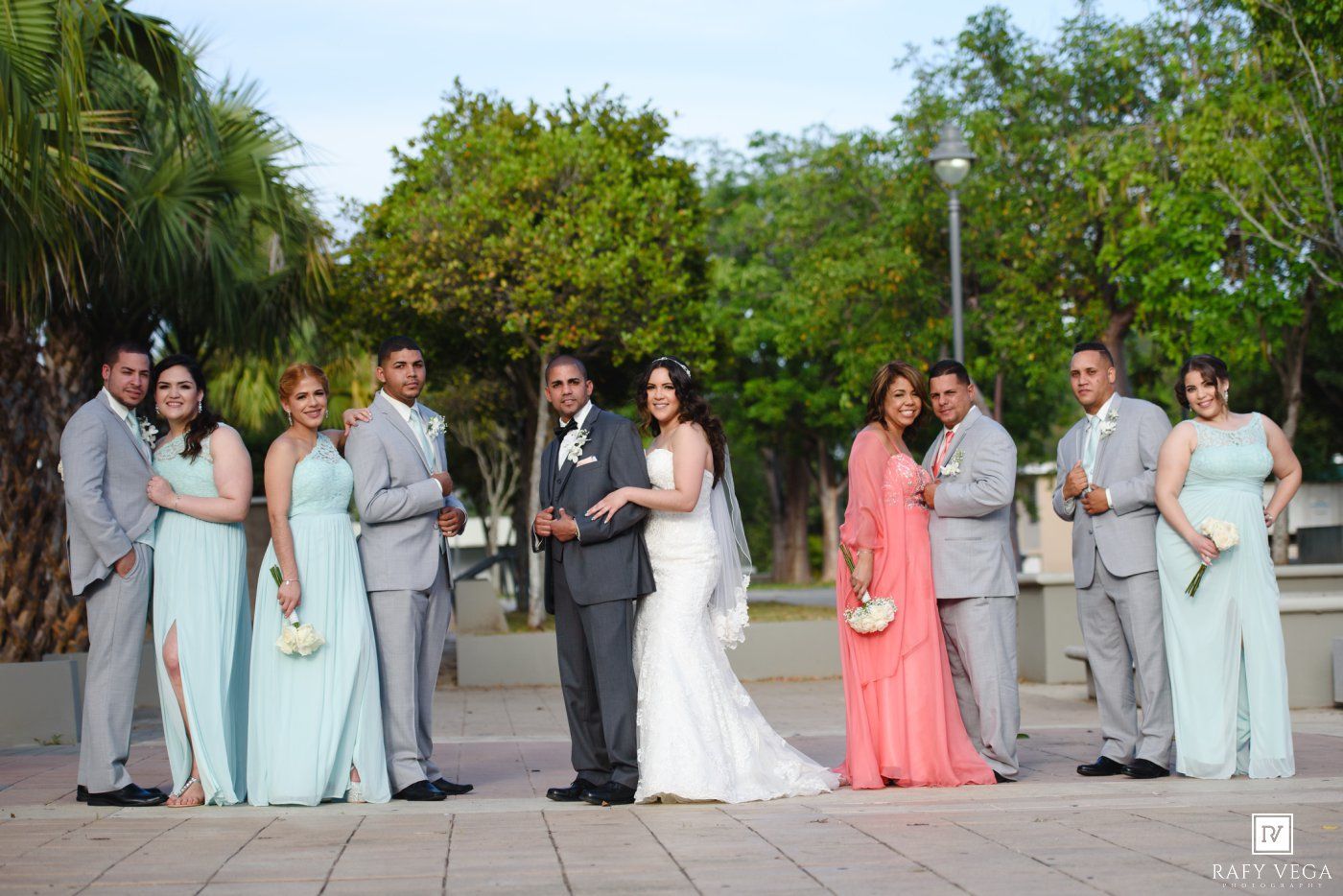 Wedding party poses outdoors with bride and groom centered, bridesmaids in mint and coral gowns, groomsmen in gray suits.