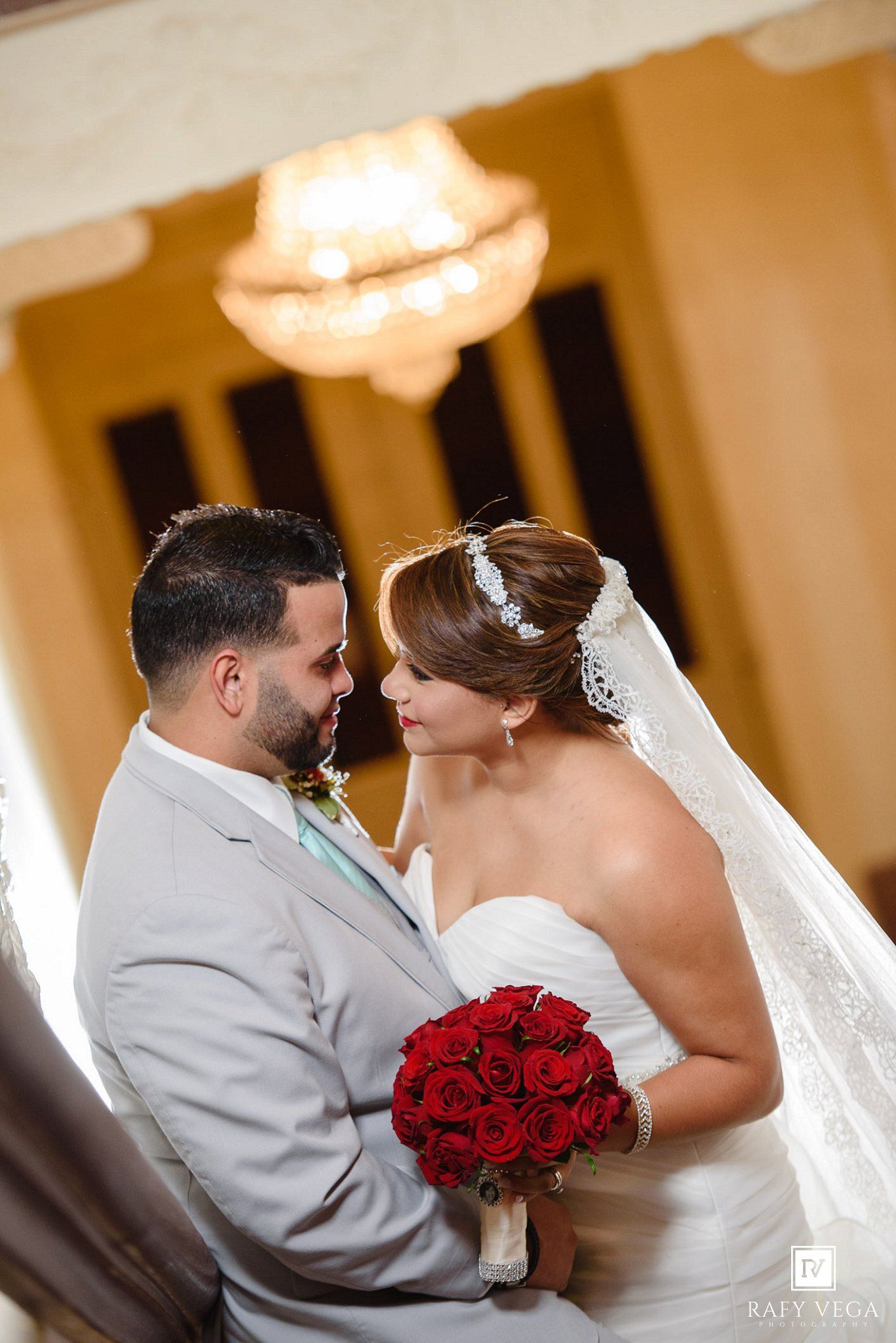 Bride and groom share a romantic moment, bride holding red rose bouquet wearing veil, groom in gray suit, elegant venue.
