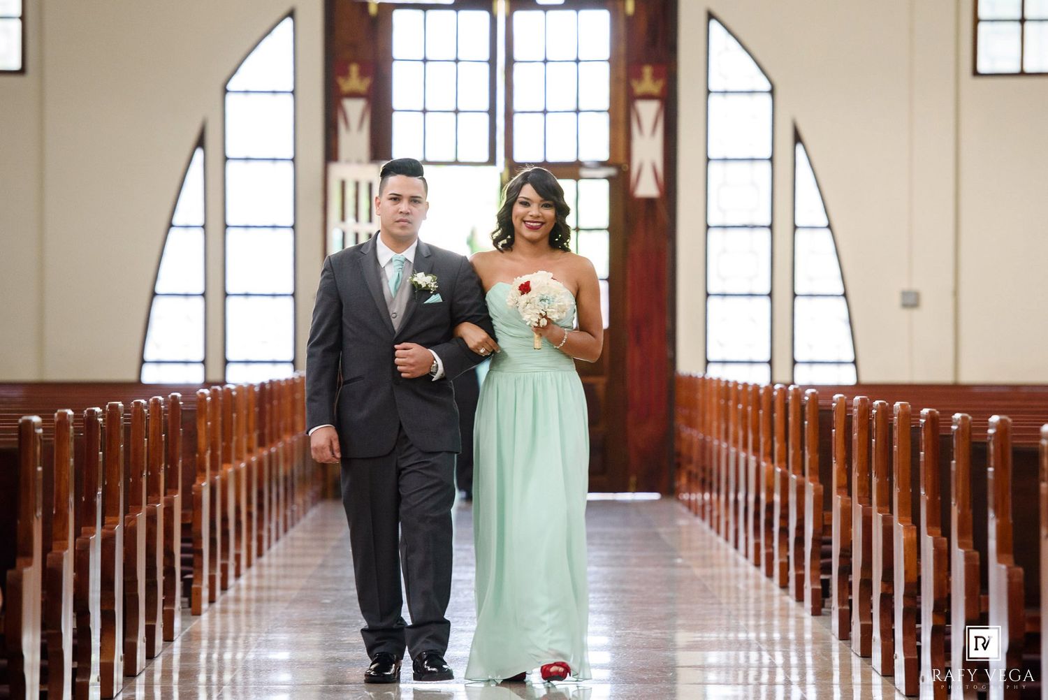 A bridesmaid in mint green gown walks down church aisle with groomsman in gray suit during wedding ceremony.