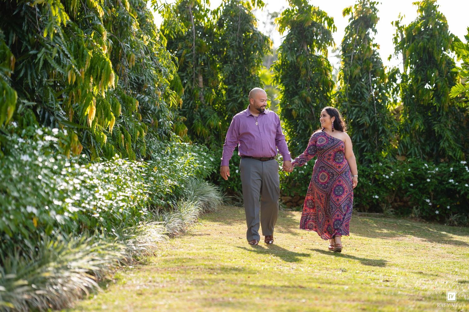 Couple holding hands walking through lush green garden path surrounded by tall trees and white flowers.