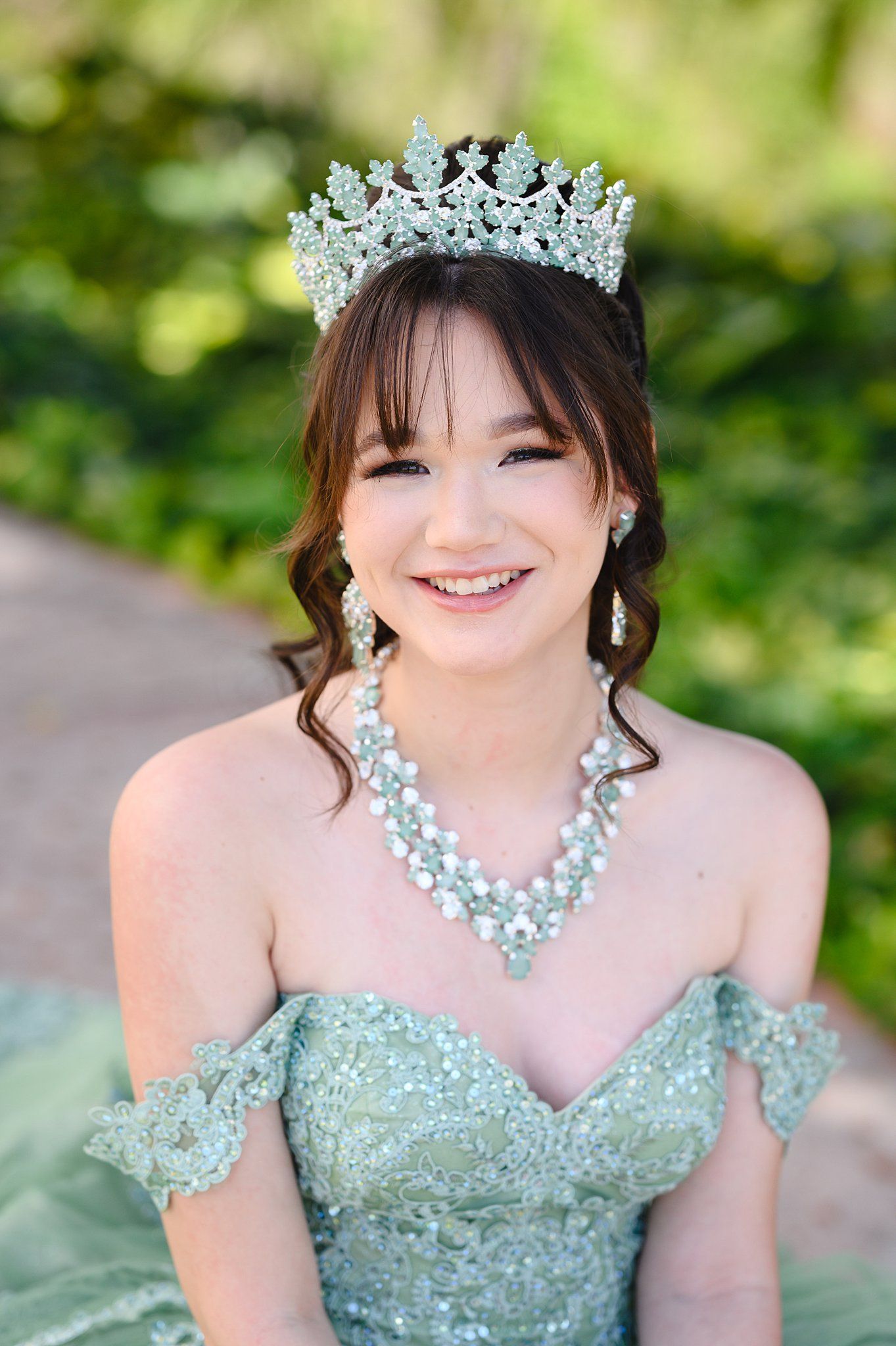 Smiling young woman in mint green off-shoulder gown with crystal tiara and pearl necklace at quinceañera celebration.