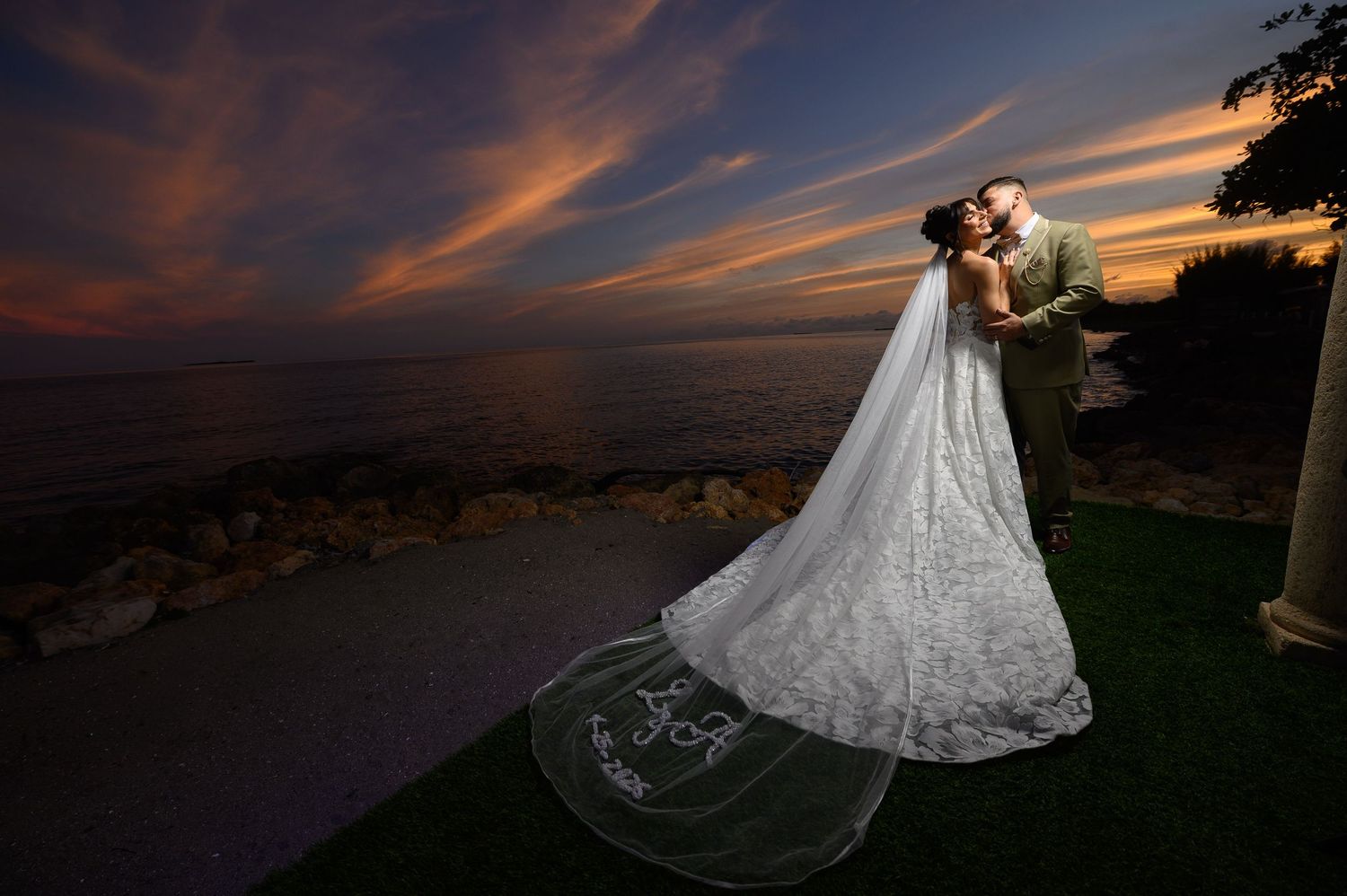Bride and groom share a romantic kiss at sunset by the ocean, her lace gown and cathedral veil trailing on green grass.