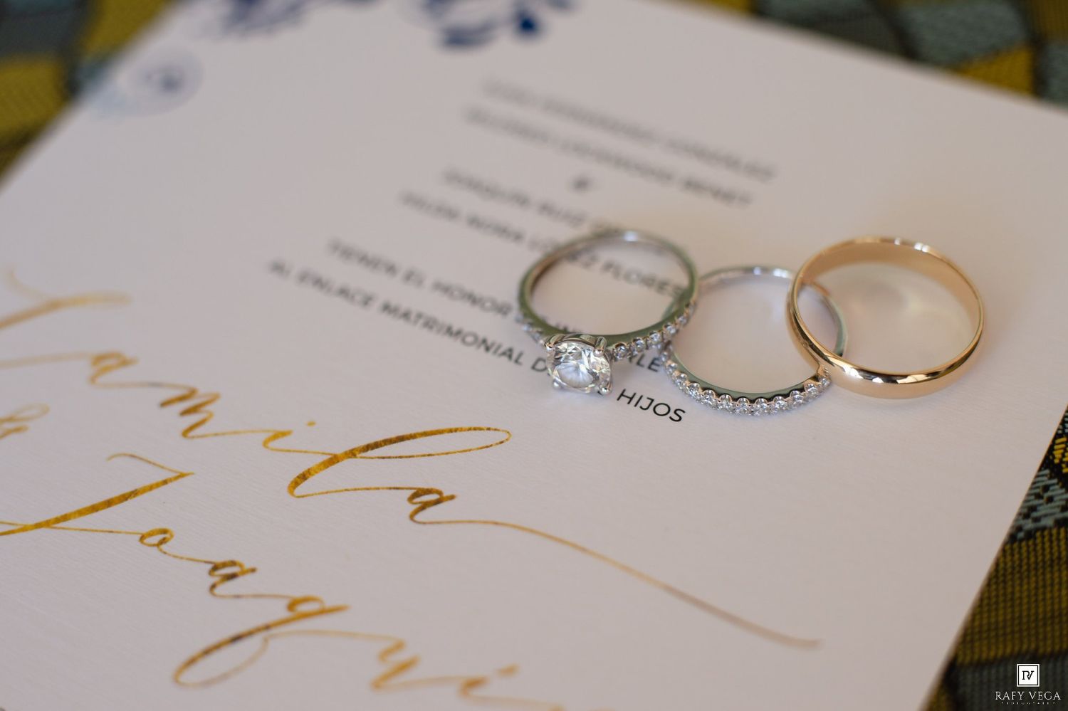 Three elegant wedding rings, including a diamond engagement ring, rest on a gold calligraphy wedding invitation.