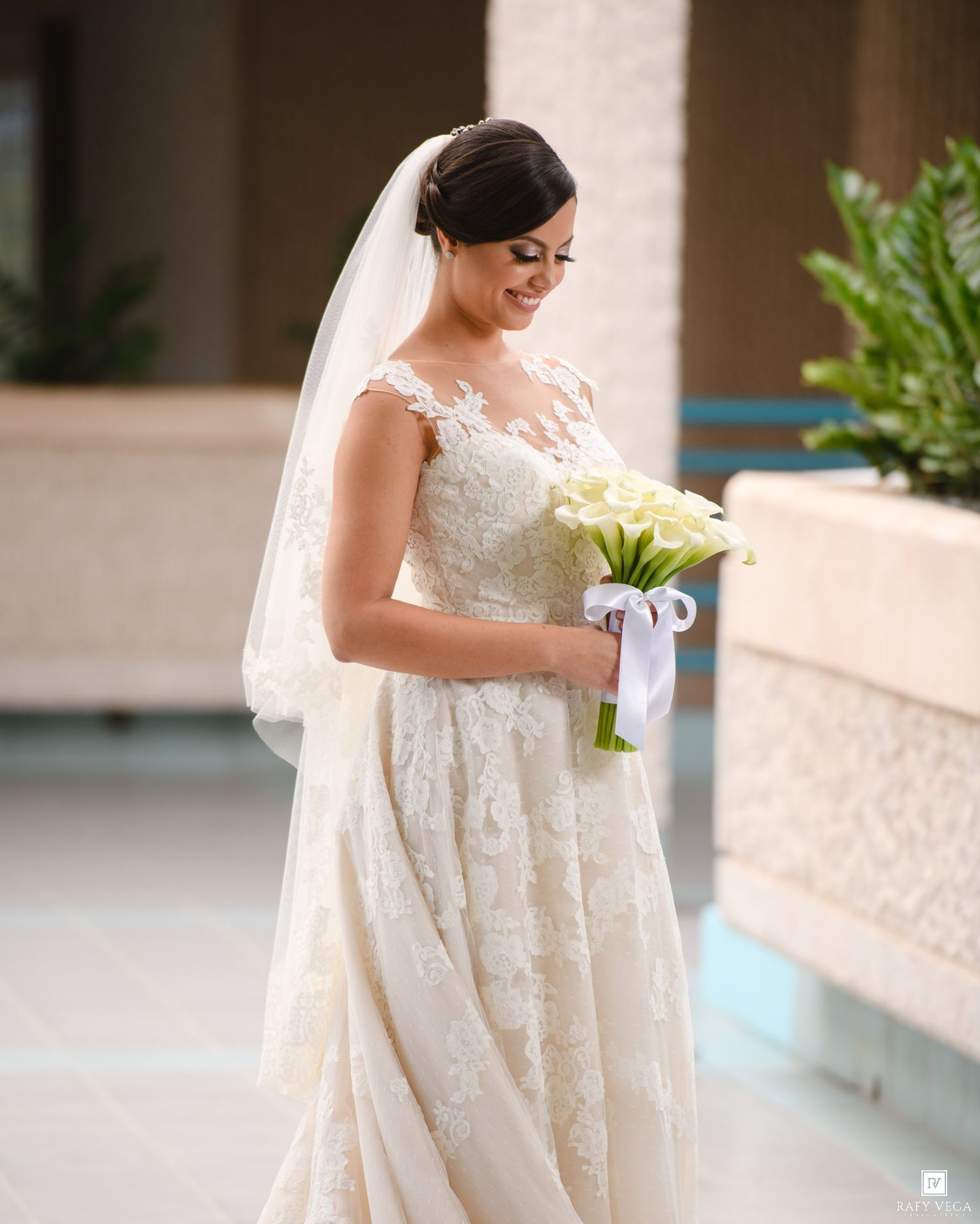 Bride in lace wedding gown holding white calla lily bouquet with veil, smiling elegantly outdoors.
