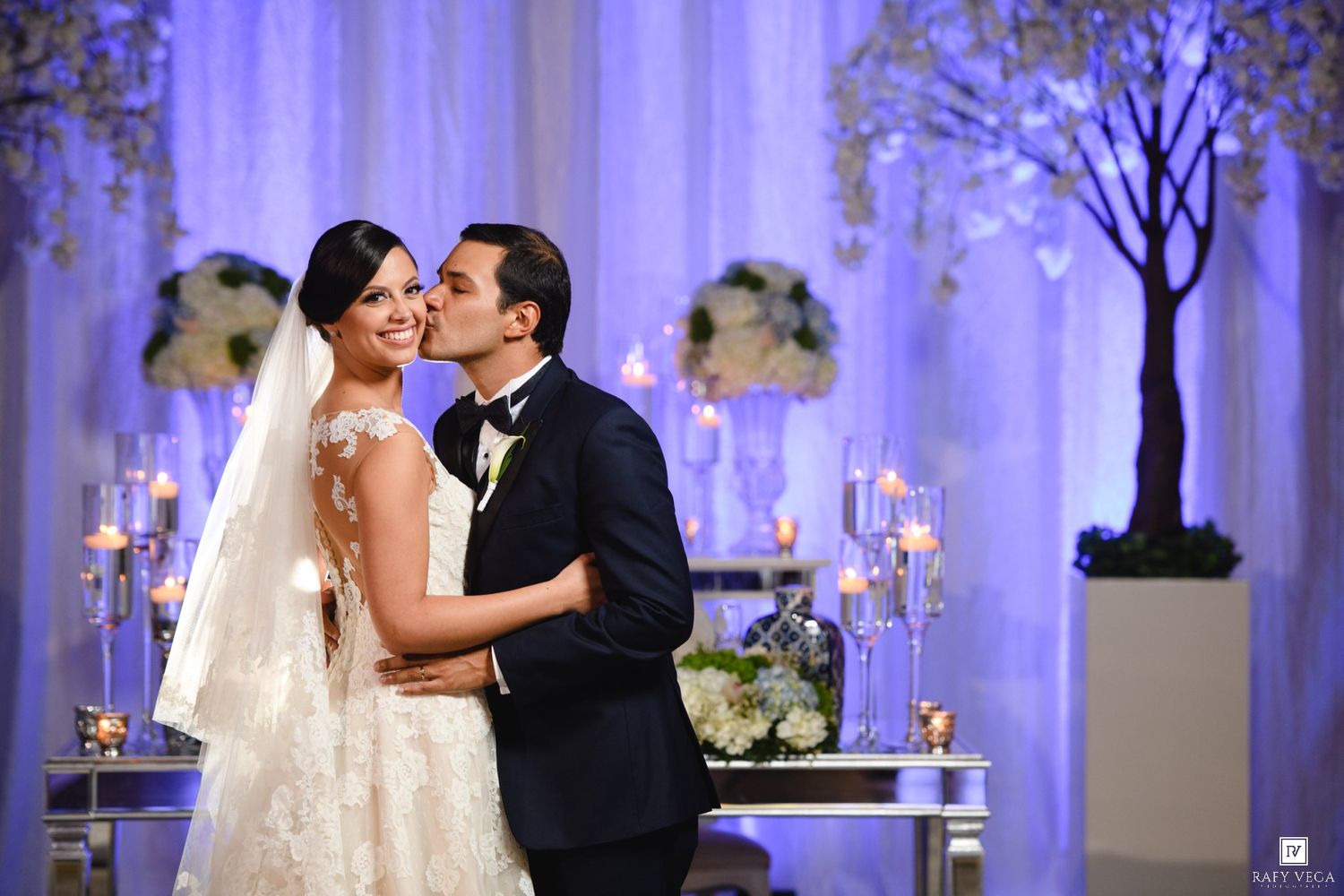 Bride and groom share a romantic kiss at elegant wedding reception with candlelit decor and white floral arrangements.
