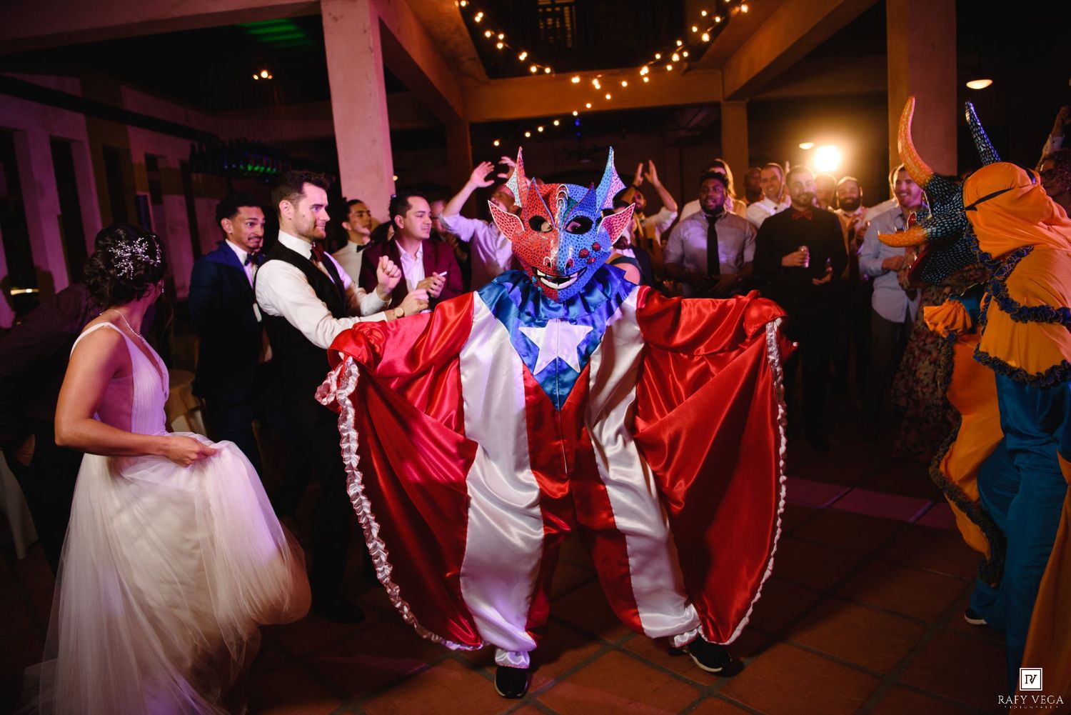 Wedding guests dancing with a colorful costumed performer in a red and white outfit at a festive reception.